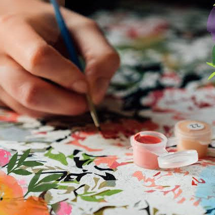 Person painting a floral paint by number design with paint pots on a table.