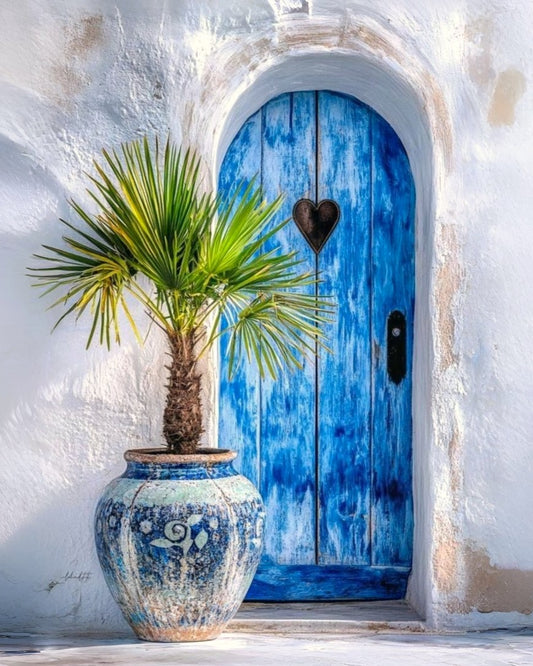 A vibrant blue wooden door with a heart-shaped metal ornament and a black handle, set in a white stucco wall. In the foreground, a potted green palm tree in a blue and white patterned ceramic pot adds to the scene.