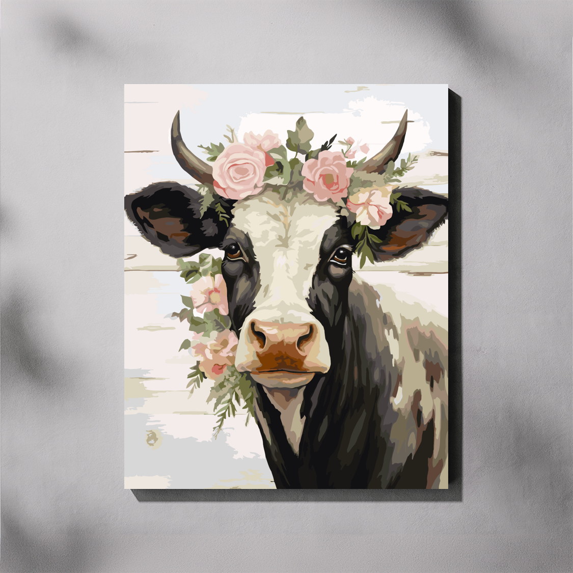 A vertical paint by number canvas featuring a painted cow with a floral crown of pink roses and green leaves, shown hanging against a light gray wall with soft shadow overlays.