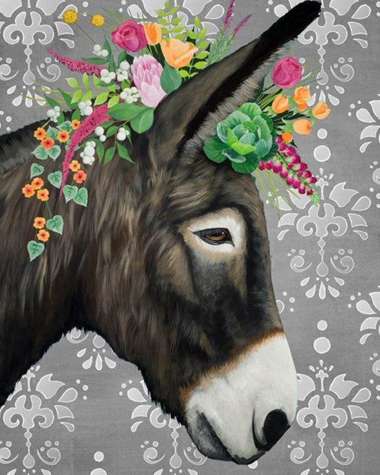 A close-up portrait of a dark brown donkey head with a white muzzle, decorated with a colorful floral crown of pink, orange, yellow, and green flowers, set against a gray damask-patterned background.