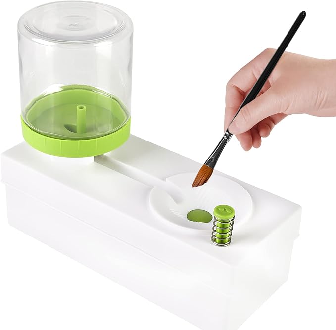Hand using a paintbrush to dip into a small container of green paint on a white tray with a clear lid.