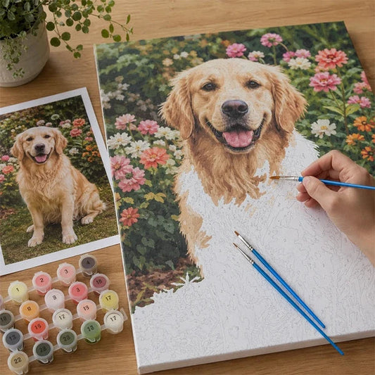 A hand is painting a custom paint by number canvas portrait of a golden retriever dog in a garden, with a printed reference photo of the dog on the left, paint tubes, and two paintbrushes on a wooden table.
