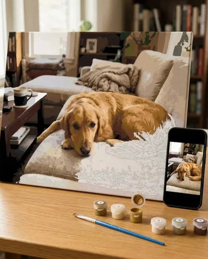 A large personalised canvas paint by number showing a golden retriever lying on a couch in a living room, displayed on a wooden table next to a smartphone showing the reference photo, with paint pots, a paintbrush, and a pencil in front.
