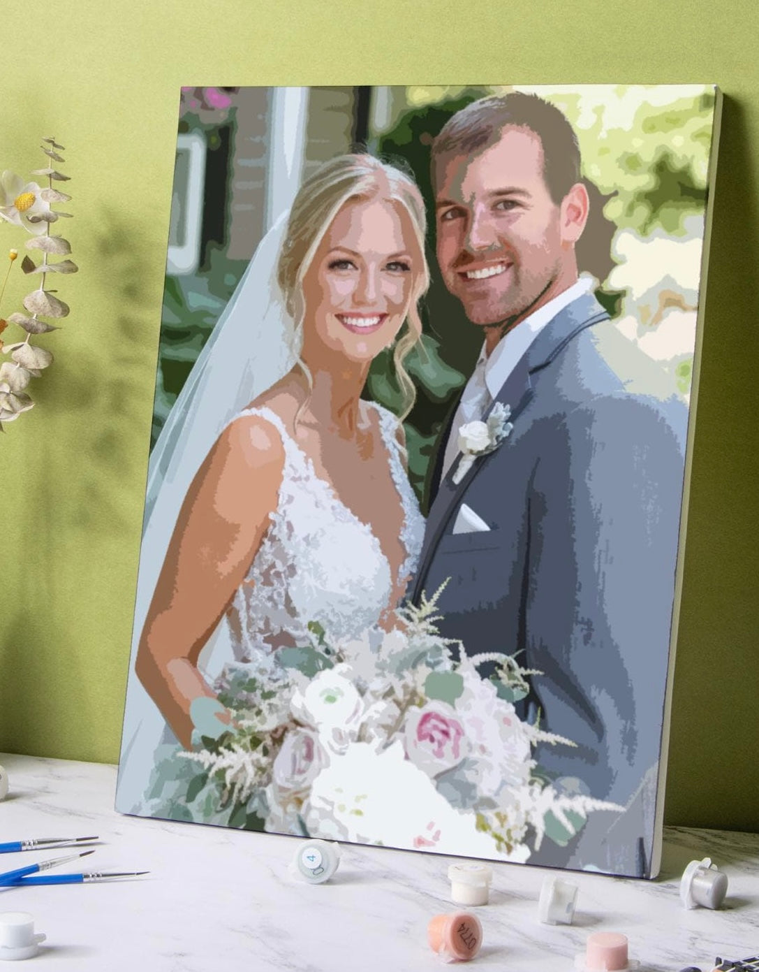 A customised paint by number canvas of a smiling bride and groom, styled on a marble tabletop with paintbrushes and paint tubes against a lime green wall.