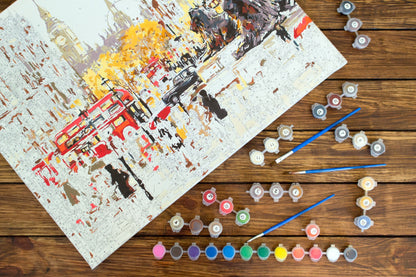 Artistic depiction of a city scene with vehicles on paper, surrounded by paint tubes and brushes on a wooden surface.